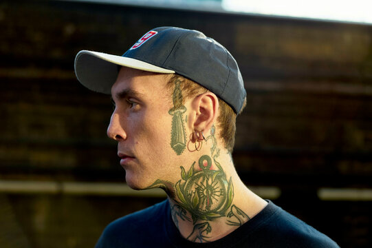 Side profile of a man with tattoos on his neck and face, wearing a baseball cap at sunset.