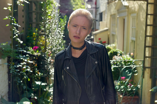 A woman in a black leather jacket showing a confused expression while standing outdoors surrounded by green foliage.