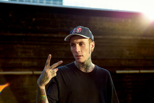 A tattooed young man in a black T-shirt and baseball cap flashes a peace sign against a sunlit background.