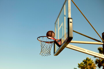 Outdoor basketball hoop with a ball going through the net against a clear blue sky.