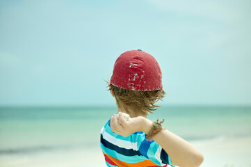 Child with a red helmet looking out at the sea on a sunny beach day.