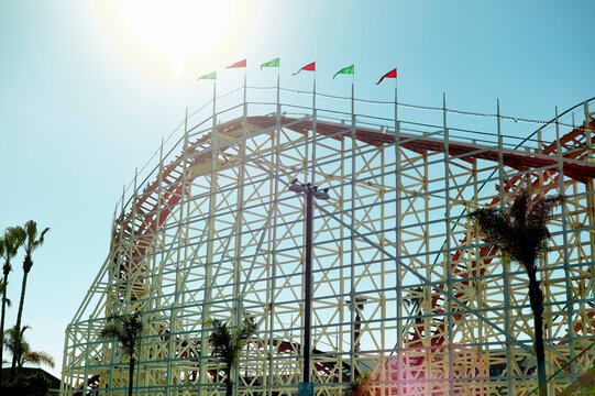 Sunlight flares over a towering wooden roller coaster adorned with colorful flags under a clear blue sky.