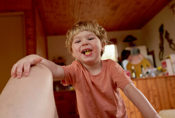 Smiling child with fair hair leaning on an adult's knee indoors, with a piece of yellow food in their mouth.