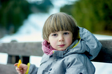 Young child sitting on a bench outdoors wearing a grey coat with a colorful scarf and holding a yellow object.