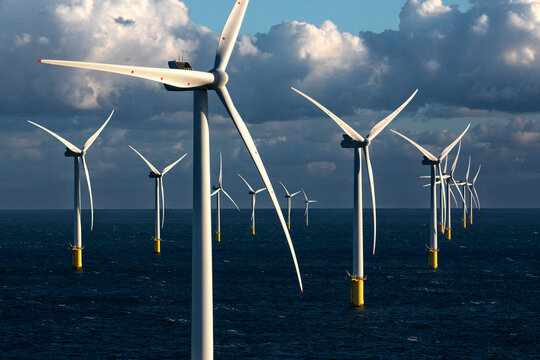 Offshore wind turbines stand tall over the ocean against a backdrop of a cloudy sky at sunset.