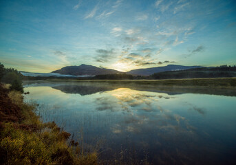 Sunrise over a tranquil lake with reflections of clouds and hills on the water surrounded by forested landscape.