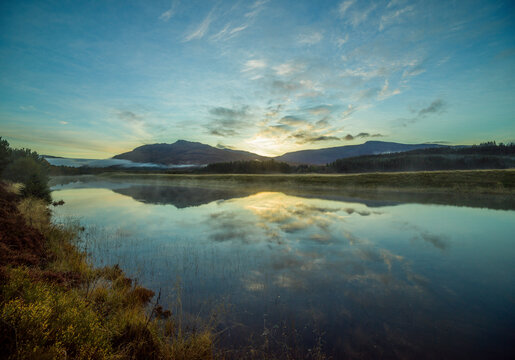 Sunrise over a tranquil lake with reflections of clouds and hills on the water surrounded by forested landscape.