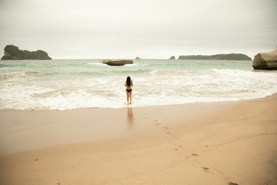 A woman stands with her back to the camera, gazing at the ocean waves and rock formations along a serene beach.
