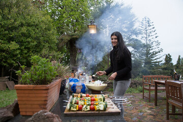 A woman grills vegetables in an outdoor setting with a smoke trail rising from the barbecue, Auckland, New Zealand