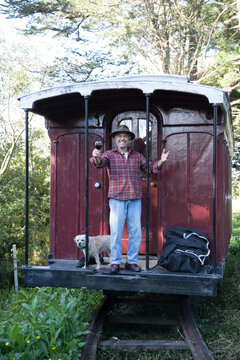 Man with a dog standing on a vintage train caboose surrounded by greenery.