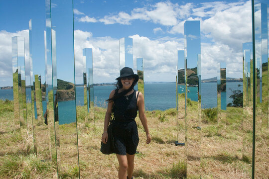 Woman in a black dress and hat standing among reflective vertical panels with a scenic coastal backdrop, Waiheke Island, New Zealand