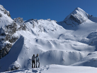 Two people stand admiring the breathtaking snow-covered mountain landscape under a clear blue sky.