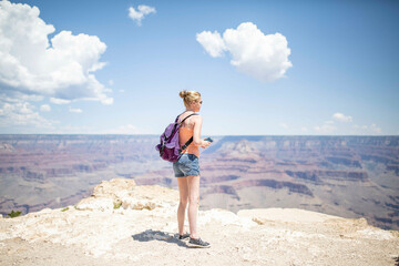 Woman standing on the edge of the Grand Canyon, looking out over the vast landscape on a sunny day with blue sky and clouds above.