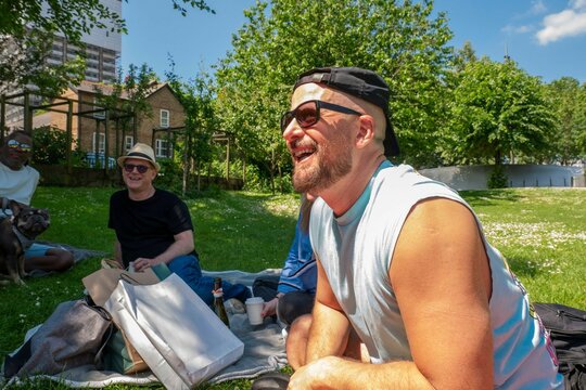 Man in sunglasses and headband laughing during a sunny park picnic with friends and a dog in the background