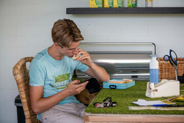 A focused young man eats lunch at a workshop bench while looking at his phone.