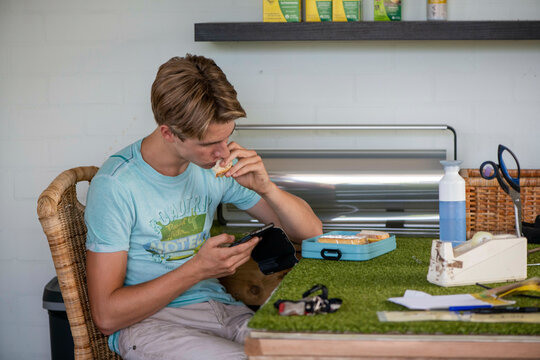 A focused young man eats lunch at a workshop bench while looking at his phone.
