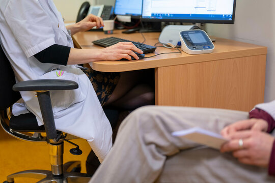 A healthcare professional is seated at a desk typing on a keyboard with a blood pressure monitor nearby, as a patient holds a document.