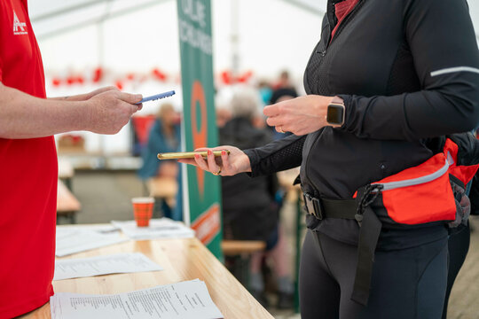A person in a red shirt hands a pen to another in a black jacket at a registration desk with banners in the background.