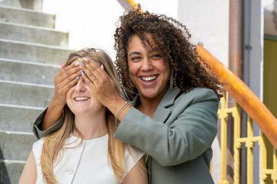 Smiling woman with curly hair covering another woman's eyes for a surprise on steps outdoors.
