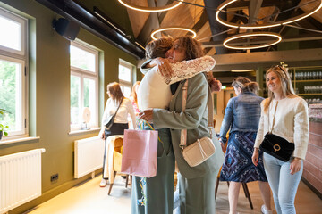 Two friends hug warmly in a cozy cafe while another woman looks on with a smile.