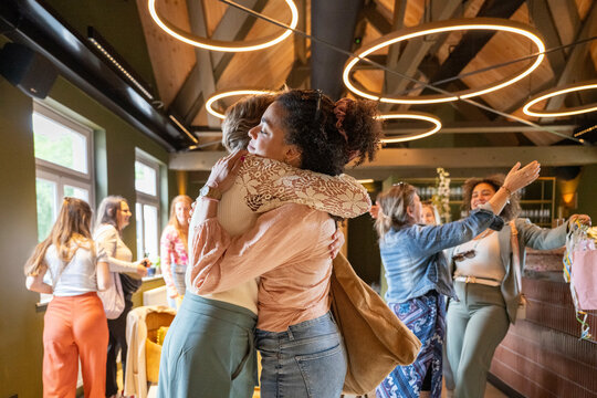 Two women embracing joyfully in a warm hug at a vibrant indoor gathering with other people chatting in the background.