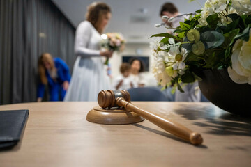 A wooden gavel rests on a table in the foreground, while a bridal party is visible in the background, inside a room that suggests a wedding ceremony.