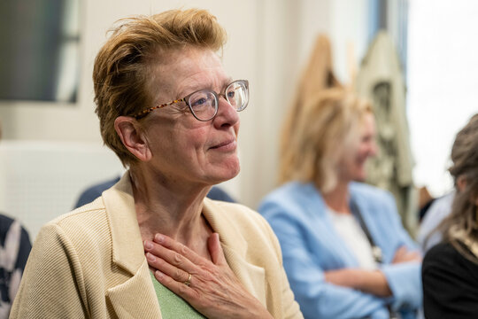 A woman with glasses wearing a beige jacket is touching her chest with a hand, showing an expression of gratitude or relief.