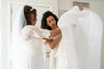 Bride in a lace wedding dress and her bridesmaid examine a wedding gown in a bright room.