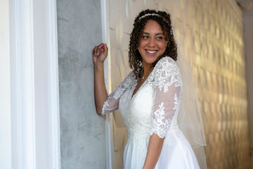 Radiant bride in a lace wedding dress with a beaming smile standing by a white textured wall.