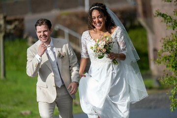 Bride in a white dress and groom in a beige suit smiling and walking outdoors on a sunny day.