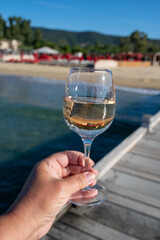 Hand with glass of cold rose wine from Provence and wooden yacht boota pier on white sandy beach Plage de Pampelonne near Saint-Tropez, summer vacation in France