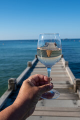 Hand with glass of cold rose wine from Provence and wooden yacht boota pier on white sandy beach Plage de Pampelonne near Saint-Tropez, summer vacation in France