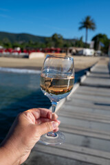 Hand with glass of cold rose wine from Provence and wooden yacht boota pier on white sandy beach Plage de Pampelonne near Saint-Tropez, summer vacation in France