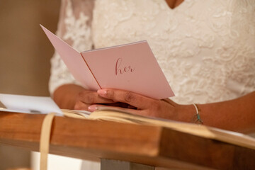 A woman in a white lace dress holds a card with 'her' written on it during a ceremony.