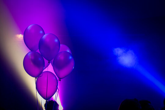 Purple balloons illuminated by blue and pink stage lights in a dark room with silhouettes of people.