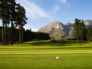 Golf ball on a lush green putting green with flagstick and dramatic mountain background under a clear blue sky.