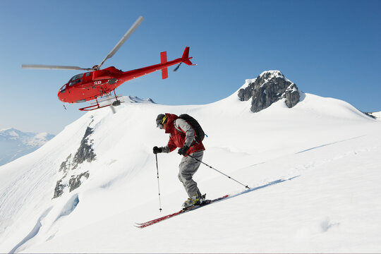 Skier in red descending a pristine mountain slope with a red helicopter in flight in the backdrop.