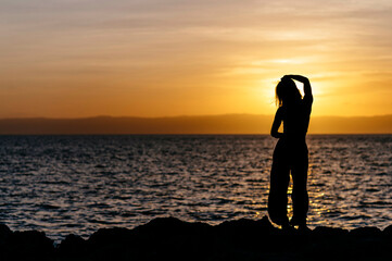 Silhouette of a person standing on rocks by the sea at sunset, with arms raised adjusting their hair.