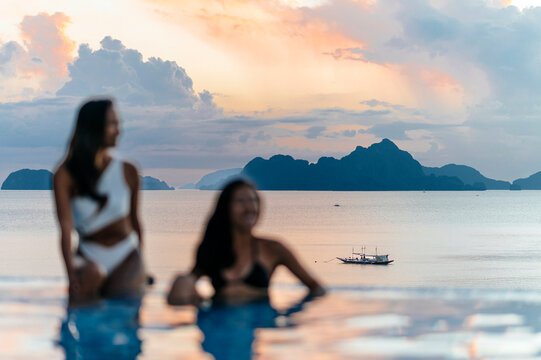 Women relax by an infinity pool with an ocean and mountain backdrop during a stunning sunset.