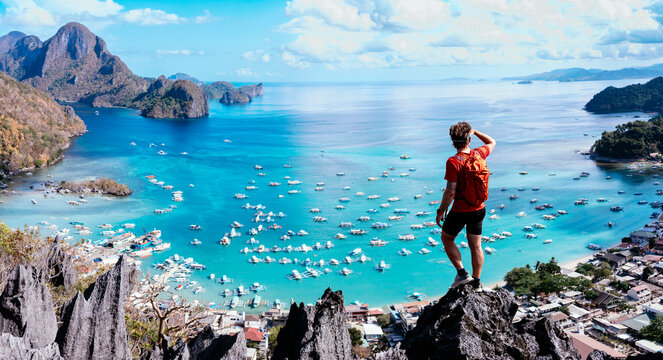 Person stands on a rocky peak overlooking a stunning blue sea scattered with boats, bordered by lush green mountains under a clear sky