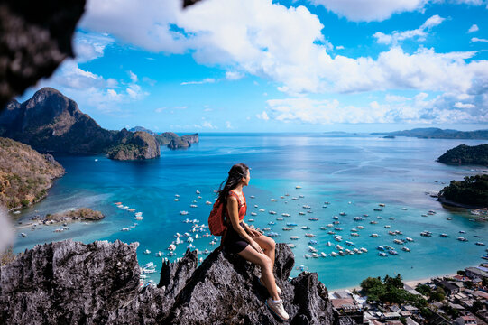A woman sits atop a rocky peak overlooking a breathtaking coastal view with boats and islands under a clear blue sky.