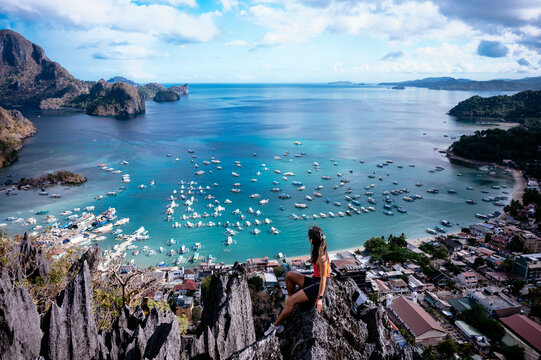 Woman sitting on a rocky peak overlooking a picturesque bay with boats and a coastal village surrounded by lush hills.
