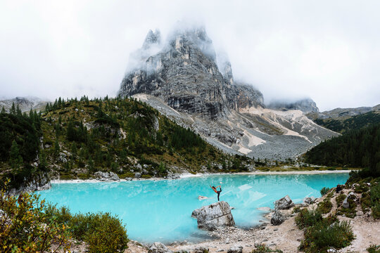 A person stands on a rock with arms raised in triumph in front of a stunning turquoise alpine lake with a majestic mountain shrouded in mist in the background.