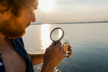 A man examines a small vial of liquid with a magnifying glass against the backdrop of a sunset over water.