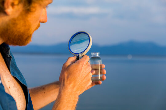 Man examining water in a small transparent bottle with a magnifying glass against a serene lake backdrop.