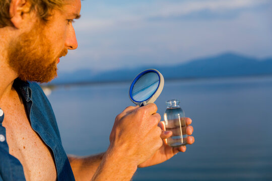 Man examining a water sample through a magnifying glass against a scenic lake backdrop.