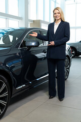 Blonde woman in elegant suit demonstrating a new car in a showroom