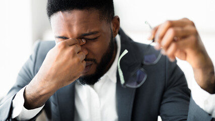 A man in a suit rubs his eyes with his hand. He holds glasses in his other hand.