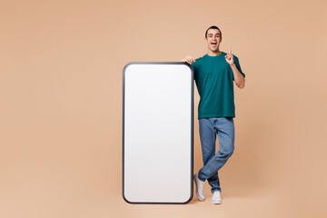 Full body young middle eastern man wears blue t-shirt casual clothes big huge blank screen mobile cell phone smartphone with workspace area point finger up isolated on plain pastel beige background.