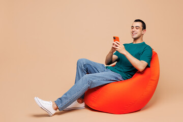 Full body young middle eastern man wears blue t-shirt casual clothes sit in bag chair hold in hand use mobile cell phone isolated on plain pastel beige background studio portrait. Lifestyle concept.